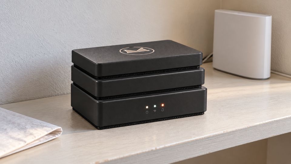 The Stack concept in Matte Black on a painted off-white shelf against a textured plaster wall, the Synkurl mark on top and three status lights glowing. A plain white router stands behind it to the right; folded white paper lies in the foreground.